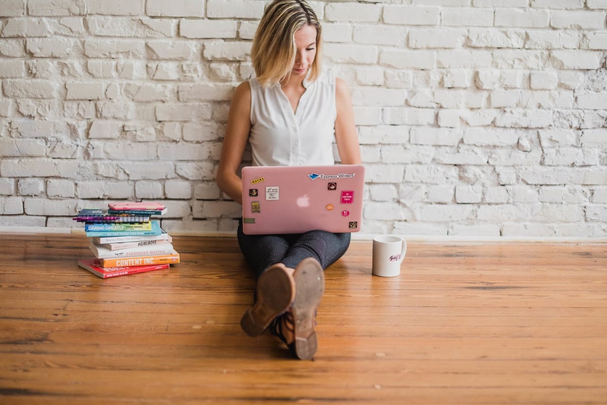 A woman with a laptop sits against a white brick wall with a cup of coffee and a stack of books.