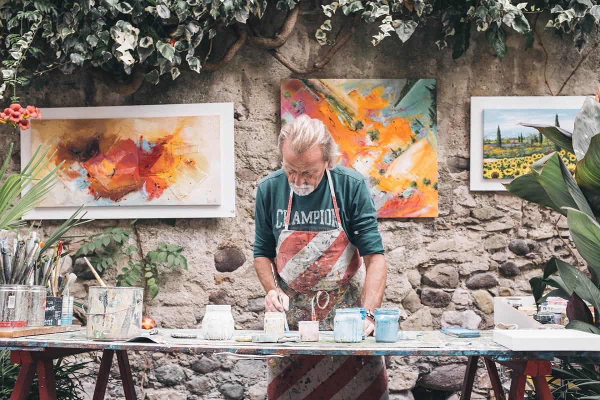 An artist painting outside with some of their work hanging behind them.